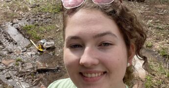female student outside hiking wearing sunglasses on head and holding up a peace sign with her fingers.